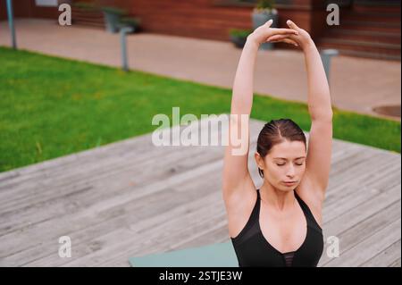 Woman doing Yoga in the Park. Woman stretching arms muscles during outdoor. Female Trainer performs arm exercises by joining the hands in a hand lock Stock Photo