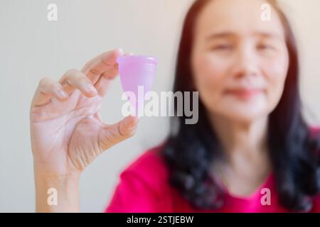 Asian woman smiling at the camera while holding a reusable menstrual cup in her hand. Selective focus on subject. Stock Photo