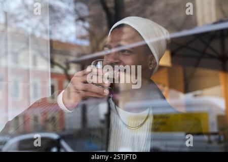 Stylish man is smiling while drinking coffee at a cafe, seen from behind a window reflecting the city street Stock Photo