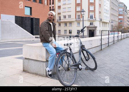 Young man takes a break on a low wall in the city, his bicycle resting beside him, enjoying the urban scenery after his commute Stock Photo