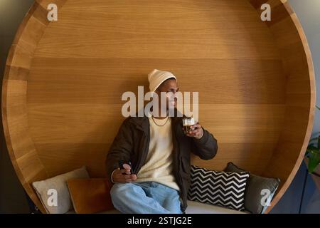Young man enjoying a beverage and using his smartphone in a trendy cafe with unique wooden seating Stock Photo