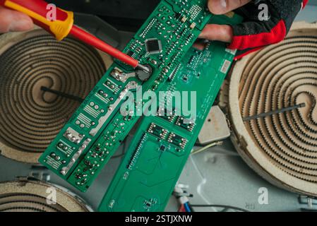Repairman using a screwdriver to fix a circuit board of a broken electric stove, performing maintenance and troubleshooting to restore functionality Stock Photo