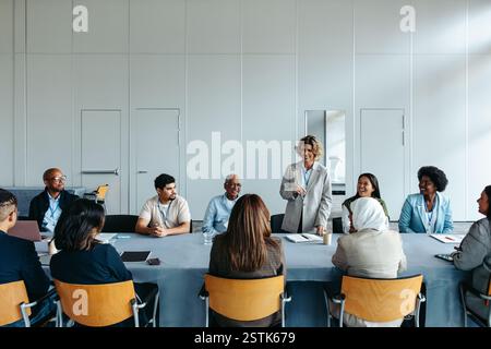 A mature female leader confidently addressing her diverse team during a business meeting in a modern office setting. Stock Photo