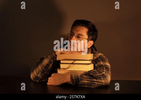 A young man in glasses and plaid shirt rests his chin on a stack of books under warm, moody lighting, reflecting a contemplative and poetic mood. Stock Photo
