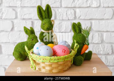 Trhee decorative green moss bunny figures with colorful Easter eggs in wicker basket on wooden table on white brick wall background. Easter holiday fe Stock Photo