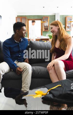 Diverse teen couple sitting on sofa, smiling and talking, graduation cap on table nearby, at home. celebration, friendship, conversation, education, m Stock Photo