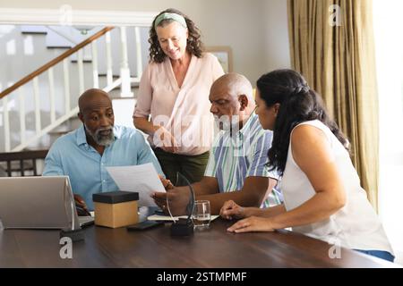 Senior friends discussing documents at home, collaborating around table with laptop. Collaboration, teamwork, discussion, planning, technology, mature Stock Photo