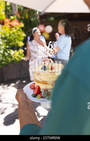 Celebrating 60th birthday outdoors with cake and joyful diverse senior friends in garden. Celebration, milestone, party, gathering, happiness, friends Stock Photo