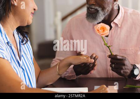Senior senior diverse couple sharing tender moment with rose, holding hands at home. Elderly, romance, togetherness, affection, love, relationship Stock Photo