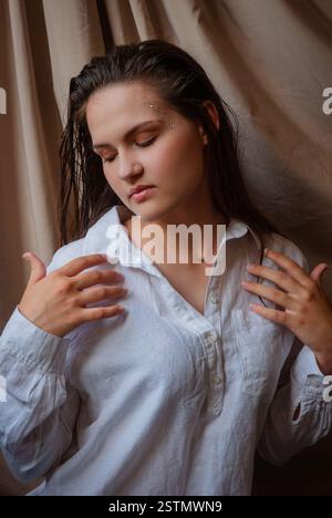 Calm contemplation with woman in white shirt against beige fabric backdrop. Caucasian woman with glitter on face. Beige background. Stock Photo