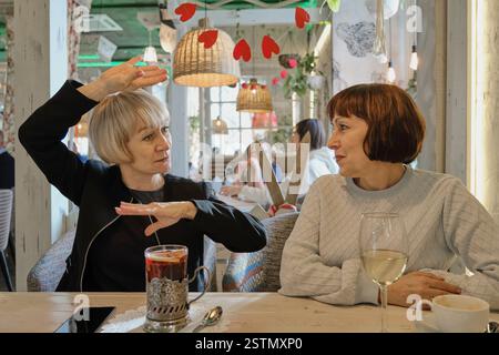 Two senior women engaged in conversation at cozy cafe. One woman in black blazer gestures expressively while other listens attentively. Warm lighting, Stock Photo