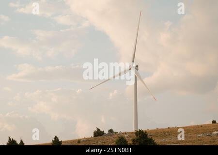 A single wind turbine stands tall against a partly cloudy sky atop a hill Stock Photo
