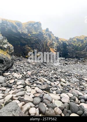 A rocky beach with smooth pebbles and steep cliffs made of dark volcanic rock, partially shrouded in mist Stock Photo