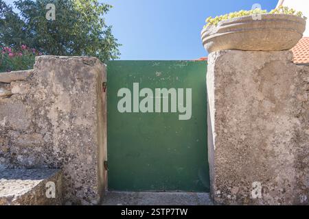 A picturesque view of a rustic green gate set in a stone wall, topped with a potted plant, under a bright blue sky. The setting conveys charm and tran Stock Photo