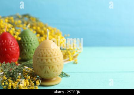 Colorful egg-shaped candles and mimosa flowers on turquoise wooden table against light blue background, closeup with space for text. Easter decor Stock Photo