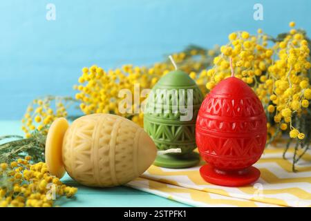 Colorful egg-shaped candles and mimosa flowers on turquoise table against light blue background, closeup. Easter decor Stock Photo