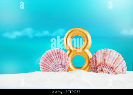 golden number 8 stands on soft white sand, by large pink seashells, against backdrop of calm blue sea on sunny day. Celebration, nature’s beauty, Inte Stock Photo