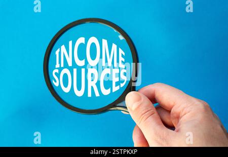 Income Sources symbol. Magnifying Glass with Concept word Income Sources. Beautiful blue background. Businessman hand. Business and Income Sources con Stock Photo