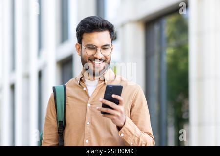 Young student with backpack on shoulder using app on smartphone. Man walking outside university campus smiling with phone in hands. Stock Photo