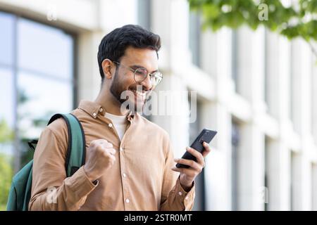 Man outside university campus with phone in hand celebrating successful exam results, student with backpack holding hand up in triumph gesture, received online notification on smartphone. Stock Photo