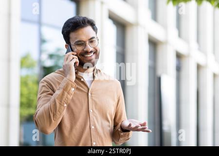 Young smiling man talking on the phone while walking outside the office building. Company employee in casual clothes, satisfied and successful programmer student. Stock Photo