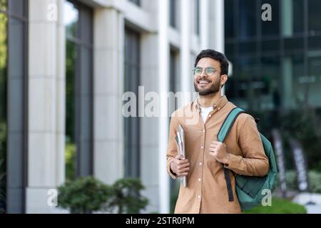 Student walking outside university campus with books in hand. Young man with backpack in casual clothes, happy and smiling. Stock Photo