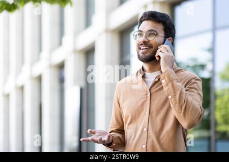 Young smiling man talking on the phone while walking outside the office building. Company employee in casual clothes, satisfied and successful programmer student. Stock Photo