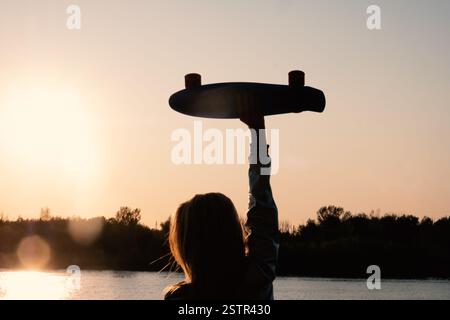 Silhouetted Woman posing with her hands holding skateboard raised up in the sunset sky Stock Photo