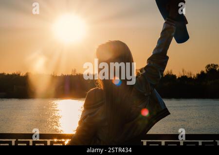 Woman posing with her hands holding skateboard raised up in the sunset sky shot with flares, low contrast image Stock Photo
