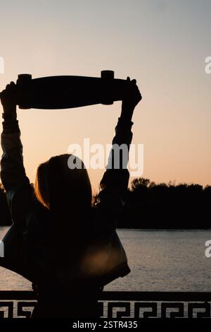 Woman posing with her hands holding skateboard raised up in the sunset sky Stock Photo