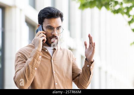 A young man with a beard wearing eyeglasses is having an argument on the phone outdoors with an angry expression and waving his hand in frustration Stock Photo