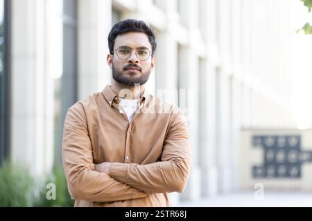 Portrait of a young bearded man with glasses standing outdoors with arms crossed. He is wearing a casual brown shirt in an urban setting Stock Photo