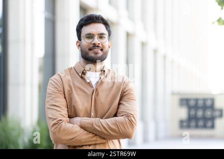 A young, attractive Indian man smiles confidently with his arms crossed in front of a modern, blurred building in the background Stock Photo