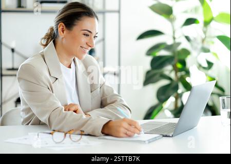 Happy pretty caucasian business lady in a suit sitting at a workspace, using her laptop and takes notes, studying information, appears focused and satisfied on the task at hand, working on a project Stock Photo