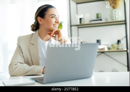 Thoughtful positive pretty caucasian businesswoman, company corporate manager or ceo, working on a laptop at a work desk while sitting in a modern office, looking away from the screen and smiling Stock Photo