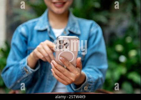 The smiling woman, with the owner face half hiding, dressed in denim jacket holding a smartphone with left hand and pointing at the screen with right Stock Photo