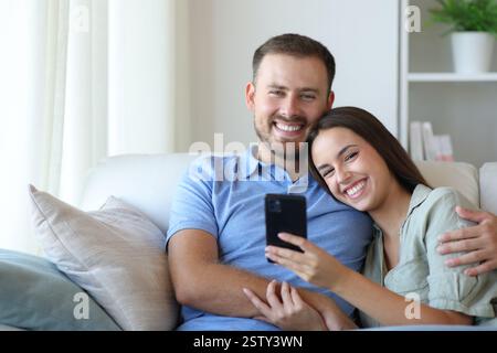 Happy couple holding phone looking at you sitting on a couch at home Stock Photo