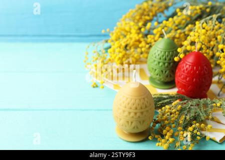 Colorful egg-shaped candles and mimosa flowers on turquoise wooden table against light blue background, closeup with space for text. Easter decor Stock Photo