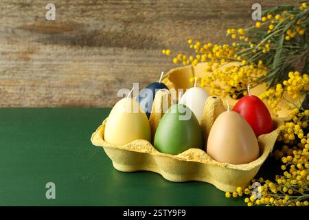Colorful egg-shaped candles in carton and mimosa flowers on green wooden table, closeup with space for text. Easter decor Stock Photo