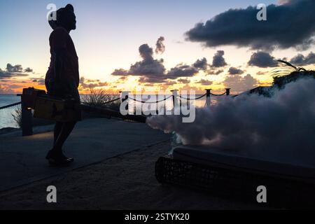 Silhouette of local man blowing thick ant-mosquito smoke against sunset sky Stock Photo