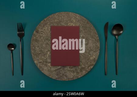 Top view of brown marble plate, black cutlery and dark red card on dark green background. Food presentation, stone tray. Table setting, copy space. Stock Photo