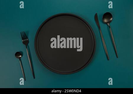 Top view of black plate and black cutlery on dark green background. Food presentation, black tray. Table setting, copy space, flat lay. Stock Photo