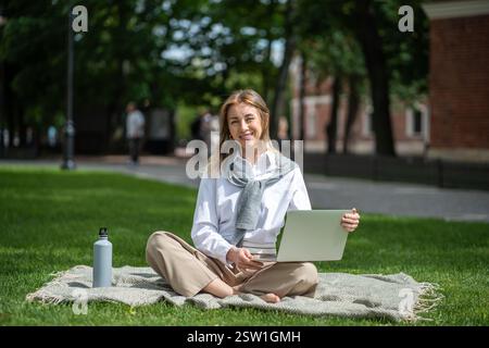 Happy businesswoman with laptop looking at camera sitting in lotus position at picnic in park. Stock Photo