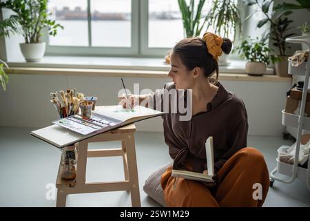 Unsmiling woman artist painting with watercolors. Female painter sitting at floor of home art studio Stock Photo