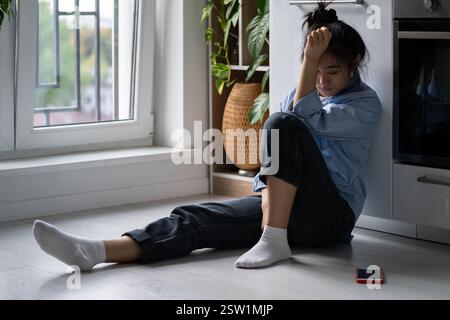 Depressed Asian girl sitting on floor at home looking at smartphone waiting for call from boyfriend Stock Photo