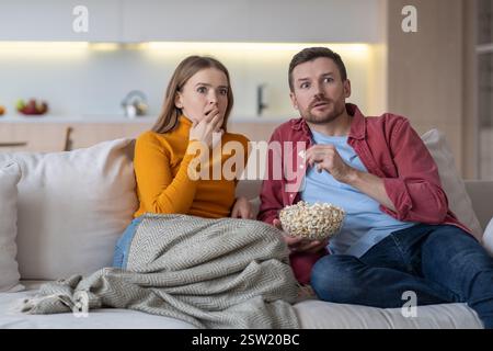 Scared married couple watching horror movie series about TV relaxing on couch, eating popcorn. Stock Photo