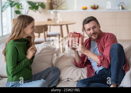 Amazed man, holding gift from loving wife, tries to guess contents of box sitting on couch at home. Stock Photo