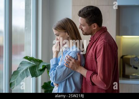 Supportive husband hugging his upset, depressive wife from behind at window in bright apartment Stock Photo