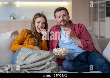 Cheerful married couple, spouses watching funny movie, series relaxing on couch, eating popcorn. Stock Photo