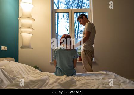 Family support. Sad woman sits on bed near standing caring loving man husband touching wife shoulder Stock Photo